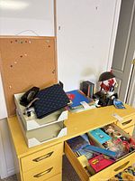 Yellow desk with cork board, desk lamp, letter trays, stapler, binder, open drawer showing assorted stationery