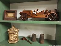 Shelf view showing framed bridge block, wooden model car, carved jar, two weights and pipe