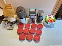 Group shot showing all items: popcorn popper, grinder, Stanley thermos with box, Rubbermaid sealed container set, twelve red-lidded jars