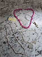 Overview of 5 necklaces laid flat: pink bead, black cord with pendant, fine gold chain, beige rope beads, silver hammered disc bib