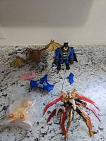 All items laid out: Batman figure, Man-Bat figure, blue accessory, bagged yellow vehicle, gold winged figure