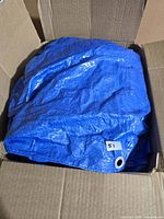 Folded blue tarp in cardboard box, grommet and lot sticker visible