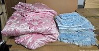 Pink satin embroidered bedspread folded next to blue fringed bedspread on table