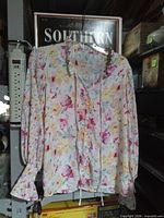 Front view of multicolor floral long-sleeve blouse on hanger showing button front, ruffle collar and drawstring ties