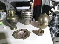 All five brass items grouped on table: lidded urn, striped vase, ribbed vase, scalloped dish, perforated cup