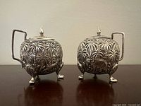 Front view of both silver condiment pots showing relief decoration and feet