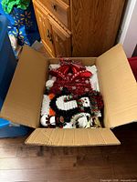 Open box displaying assorted Christmas decorations including red metallic bow, tinsel Santa hat and tinsel snowman