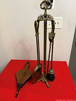 Brass fireplace tool set with stand, shovel, poker, brush, and separate wooden bellows on red surface