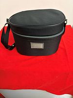 Front view of black oval makeup travel bag showing strap, lid and logo patch