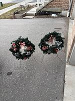 Two decorated artificial Christmas wreaths on black metal easel stands positioned on driveway