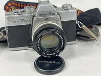 Front view of Minolta SRT 100 with attached 55mm lens, lens cap and strap visible