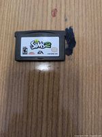 Front view of The Sims 2 GBA cartridge on wood surface