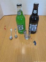Two Route 66 soda bottles, two teaspoons, one shot glass on tabletop