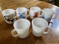 Six white milk glass mugs, four with printed graphics, two plain