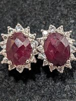 Front view of the two silver earrings showing oval dyed ruby centers and halo of clear stones