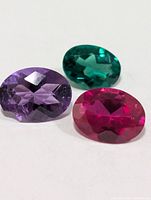 Three faceted oval gemstones in purple, green and red-pink on white background