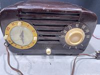 Front view of brown bakelite clock radio showing clock face, tuning dial, knobs, grille and power cord