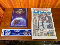 Overview of three 1992 Blue Jays World Series souvenir items: scorebook, printed cloth front page, bumper sticker