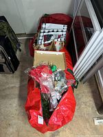Open red storage bag containing artificial tree sections, box with ornament bags and folded fabrics