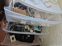 Three open plastic drawers filled with cables, adapters, camera, pouch