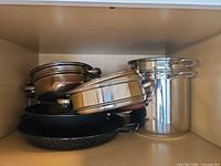 Stacked stainless steel pots, black nonstick pan and speckled enamel pan inside cabinet