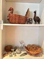 Overall view of lot on shelving showing wicker basket, wooden figurines, painted bowl, glass rabbit, boxes, arrowhead