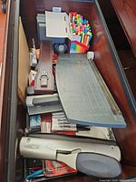 Drawer view showing paper trimmer, hole punches, markers, slide rule case and assorted supplies