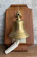 Front view of brass bell mounted on wood plaque with rope pull and engraved plate visible