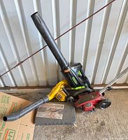 All four items leaning against wall: Greenworks blower, Paramount blower, Craftsman edger, kneeling pad