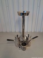 Front view of all silver plated items: two coffee pots, pedestal, cup, battery for scale