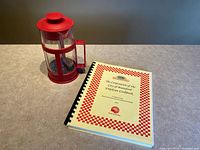Red French press beside yellow spiral-bound Brantford employee cookbook.