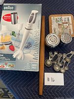 All lot items arranged: Braun hand blender box, wooden rolling pin, stainless containers, measuring spoons, trivet