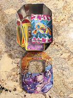 Open tin showing Mewtwo lid, tin base, loose cards, sealed booster