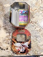 Open metal tin with stack of Pokémon cards and separate lid featuring Reshiram artwork