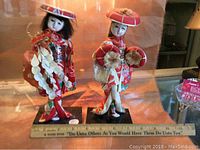 Front view of two Oriental dolls in traditional red costumes and hats with flowers, standing on black labeled bases with a ruler for scale.
