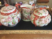 Photo showing the three hand painted porcelain items: sugar bowl with lid, lidded bowl or pot, and pitcher, all decorated with Asian motifs and floral patterns. Ruler included for scale.