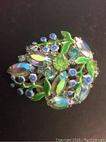 Close-up of vintage brooch with blue and green stones and enamel leaves showing floral design.