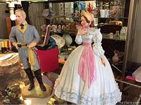 Two porcelain figurines depicting Ashley and Melanie Wilkes from Gone With The Wind, standing on reflective surface with household background.