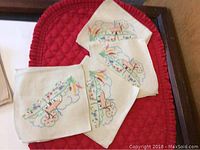 Four cream-colored napkins with colorful embroidered pastoral scenes featuring houses, trees, and clouds, folded and arranged on red quilted surface.