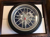 Front view of large wall clock resembling a car tire with silver rim and visible clock face.