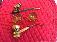 Two smoking pipes with one having a wooden and brass construction and one having a light-colored brass overlay on the bowl and an amber stem, alongside two small amber glass beer mugs.