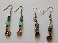 Photo showing two pairs of earrings side by side on a white background, clear view of stones and metal charms.