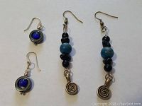 Two pairs of earrings on white background: one pair of dangle earrings with blue and black beads, spiral charm; single blue stone circular earrings