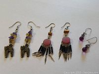 Photo showing three pairs of earrings: brass cats with stones, pink aboriginal dangles, and amethyst drops arranged flat on white background