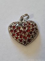 Front view of small heart-shaped pendant with many small red stones embedded in silver-tone metal.
