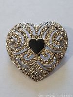 Front view of the heart-shaped pin showing detailed filigree metal openwork with sparkling bits and small solid black heart in the center.