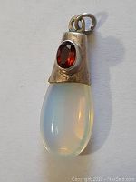 Image of the pendant showing the clear opalescent teardrop stone with red faceted gemstone inset in silver-tone metal cap, displaying iridescence and attached loop.