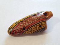 Close-up image of a small ceramic whistle decorated with Aboriginal-inspired patterns in yellow, red, and black. Shows the shape, finger holes, and mouthpiece clearly.
