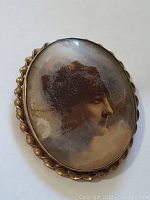 Front image of the oval brooch showing the hand painted female portrait, gold toned twisted rope frame, and aged finish.