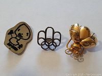 Front view of three pins: cartoon humorist character, Olympic rings, gold-toned fly